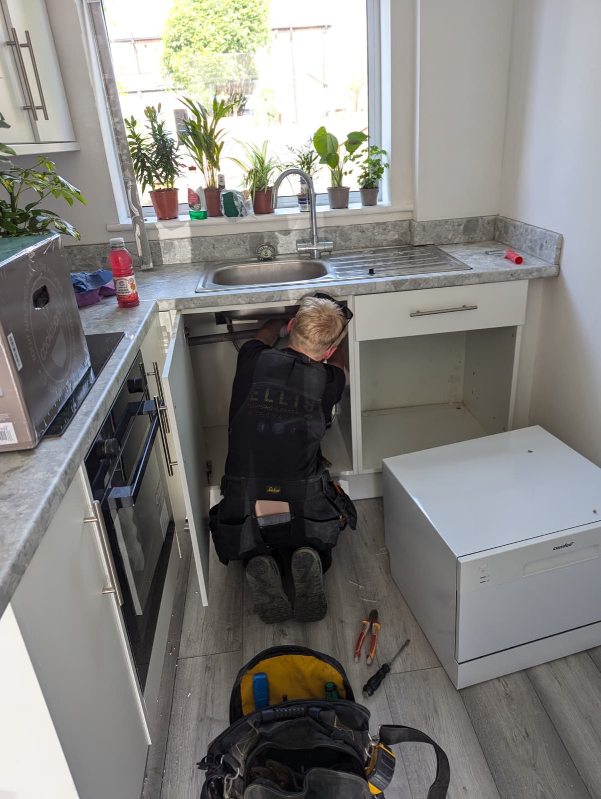 A plumber at work under the kitchen sink during a refurb, plants lining the windowsill